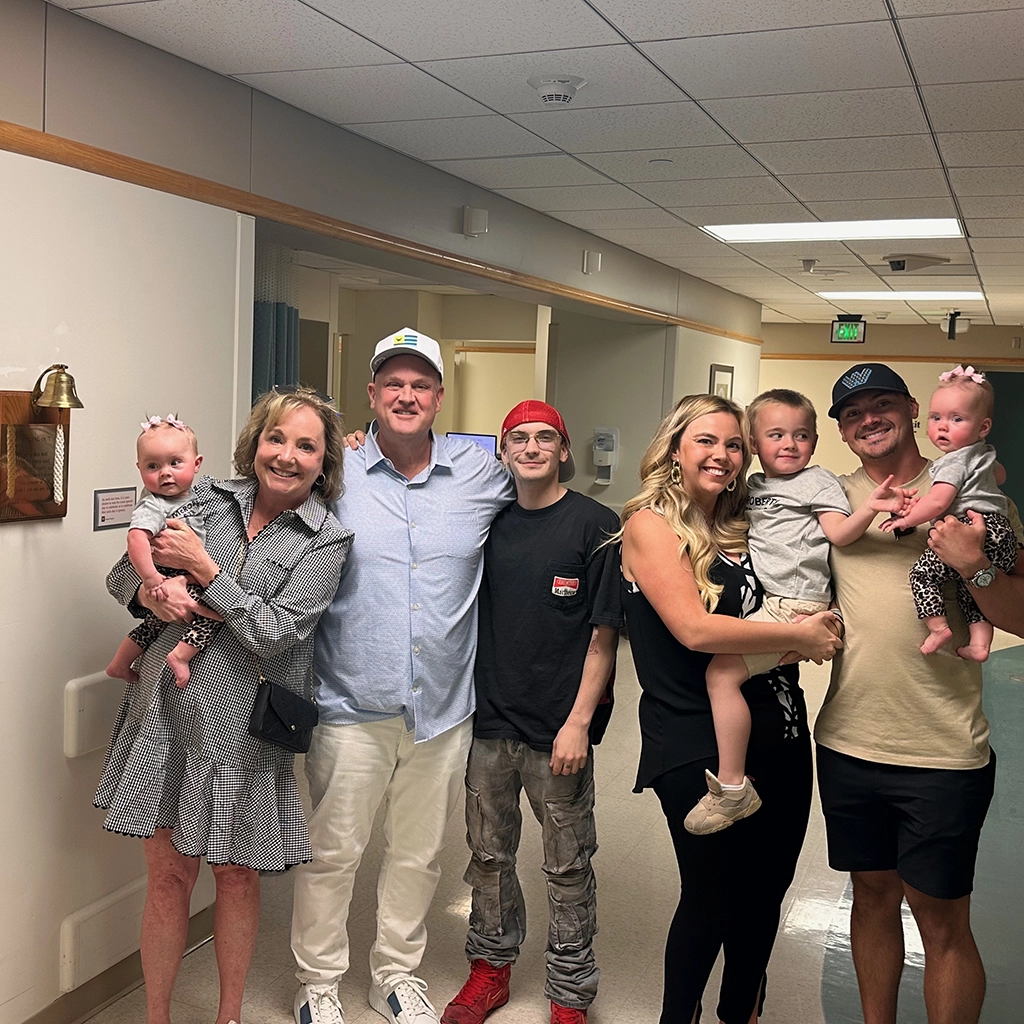 A group of six adults and three babies pose together in a hospital hallway, smiling at the camera.