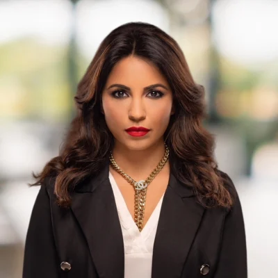 A woman with long brown hair, wearing a black blazer, white blouse, bold red lipstick, and a gold chain necklace, stands in front of a blurred outdoor background.