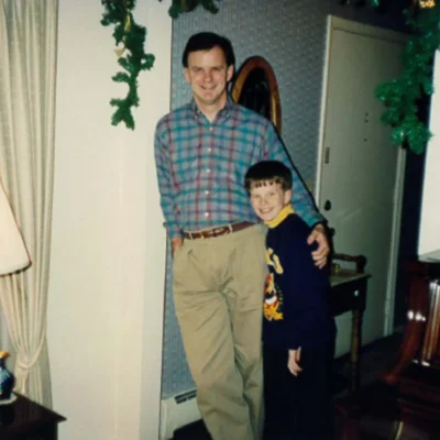 An adult and a child stand together in a living room decorated with green garlands, near a lamp and piano, both smiling at the camera.