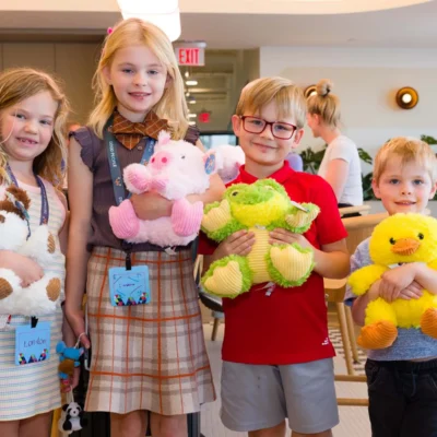 Four young children stand indoors holding stuffed animals and smiling at the camera; colorful balloons and tables are visible in the background.