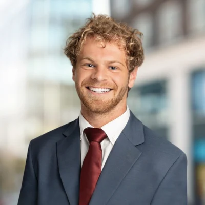 A young man with curly blond hair and a beard smiles, wearing a gray suit and red tie, standing in front of a blurred office building background.