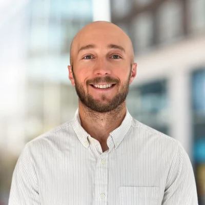 A man with a shaved head and beard, wearing a light-colored striped button-up shirt, stands smiling in front of a blurred urban background.