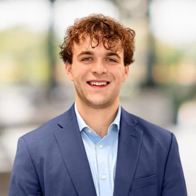 A young man with curly brown hair, wearing a blue suit jacket and light blue shirt, smiles at the camera with a blurred background.
