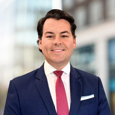 Man in a navy suit, white shirt, and red tie smiling at the camera, standing in front of a blurred office building background.
