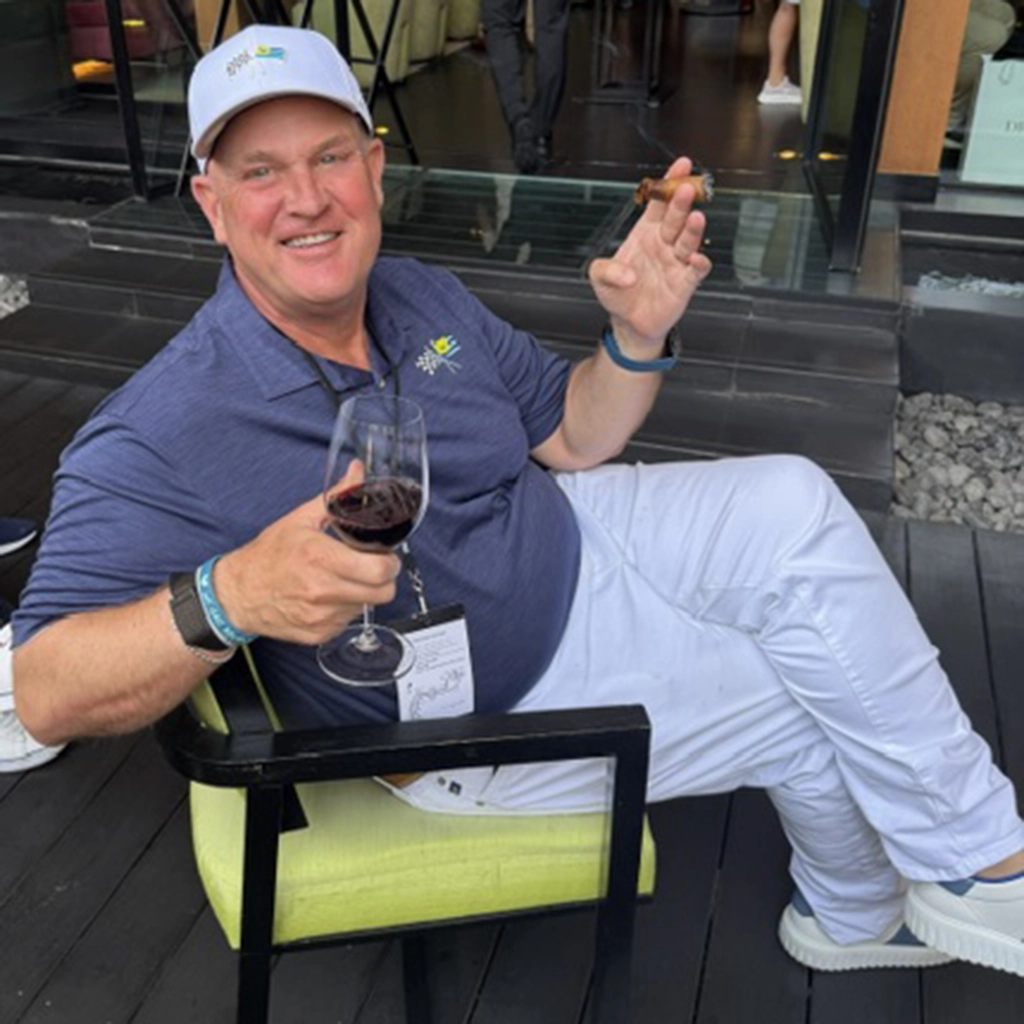 A man in a blue shirt and white pants sits on a green chair, holding a glass of red wine and a cigar, smiling at the camera.