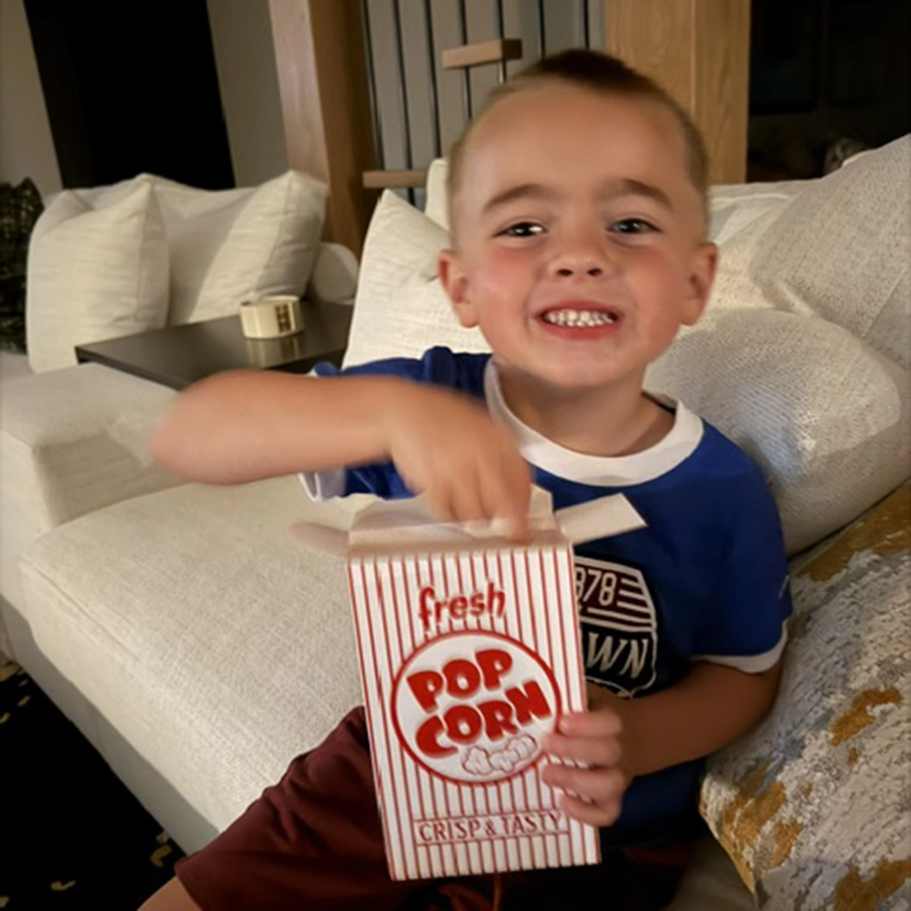 A young boy sits on a couch, smiling and reaching into a box of popcorn with "fresh POP CORN" printed on it.