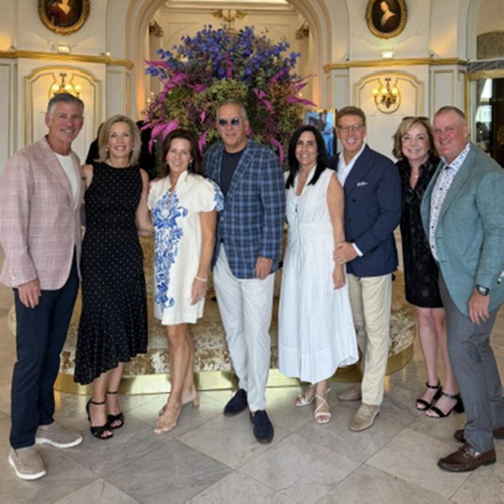 Seven adults stand together in a hotel lobby, smiling for the photo. They are dressed in semi-formal clothing, with a large floral arrangement in the background.