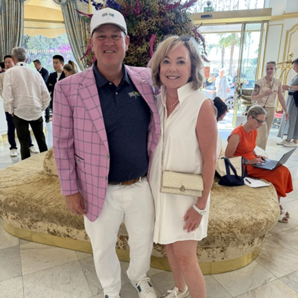 A man in a pink checked blazer and a woman in a white dress pose together in a stylish indoor setting with people in the background.