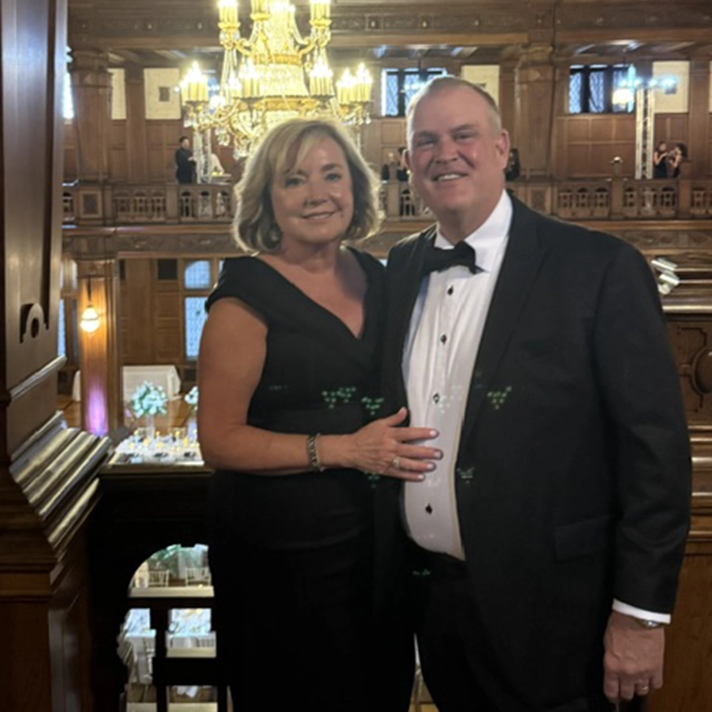 A woman in a black dress and a man in a black tuxedo stand together, smiling, in an elegant, wood-paneled hall with chandeliers and a balcony.