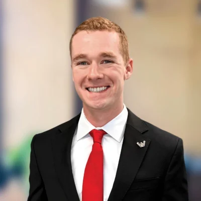 A young man in a black suit, white shirt, and red tie smiles at the camera. The background is blurred and neutral.