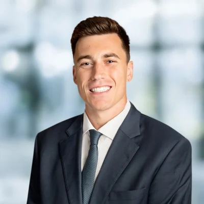 A young man in a dark suit and tie smiles at the camera against a blurred, light-colored background.