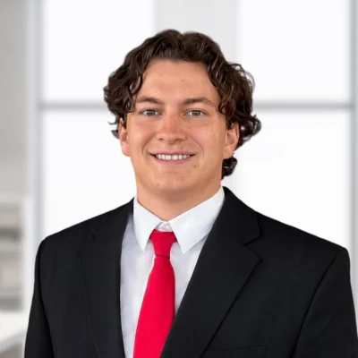 A young man with wavy brown hair wearing a black suit, white shirt, and red tie, smiling in front of a blurred office background.