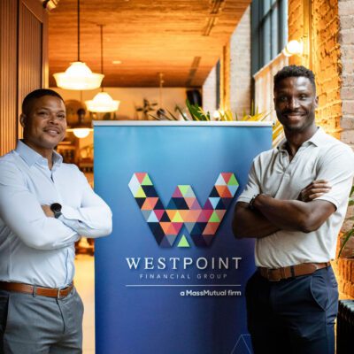 Two men stand smiling with arms crossed beside a "WestPoint Financial Group" banner in a modern, well-lit office space with brick walls and plants.