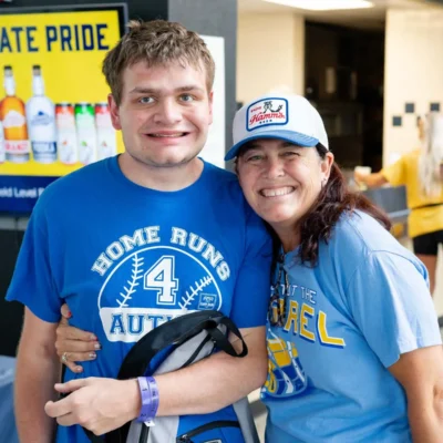 A young man and a woman smiling and posing together at an event, both wearing blue shirts, with a concession stand in the background.