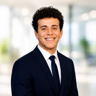 A young man with curly hair in a navy suit and tie smiles at the camera in a brightly lit office setting.