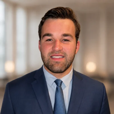 A man wearing a blue suit and tie stands indoors with a blurred background. He has short brown hair and a trimmed beard, and he is looking at the camera with a neutral expression.