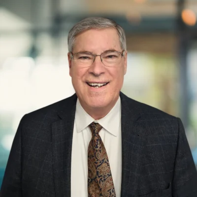 Smiling middle-aged man with gray hair, glasses, and a dark plaid suit jacket over a white shirt and patterned tie, standing indoors with a blurred background.