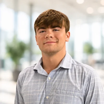 Young man with brown hair wearing a light blue plaid shirt standing indoors with a blurred background of windows and plants.