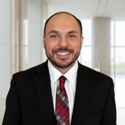 A man in a black suit, white shirt, and red patterned tie smiles while standing in a bright, modern office lobby.