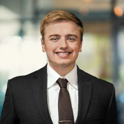 A young man in a dark suit and tie smiles at the camera, standing indoors with a blurred office background.