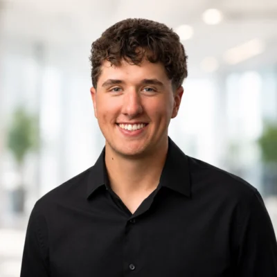 A young man with short curly brown hair, wearing a black button-up shirt, smiles at the camera in a bright indoor setting with a blurred background.