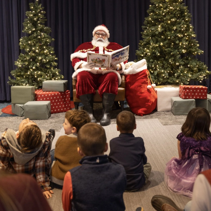 Santa Claus sits in a chair reading a book to a group of children, surrounded by Christmas trees and wrapped presents.