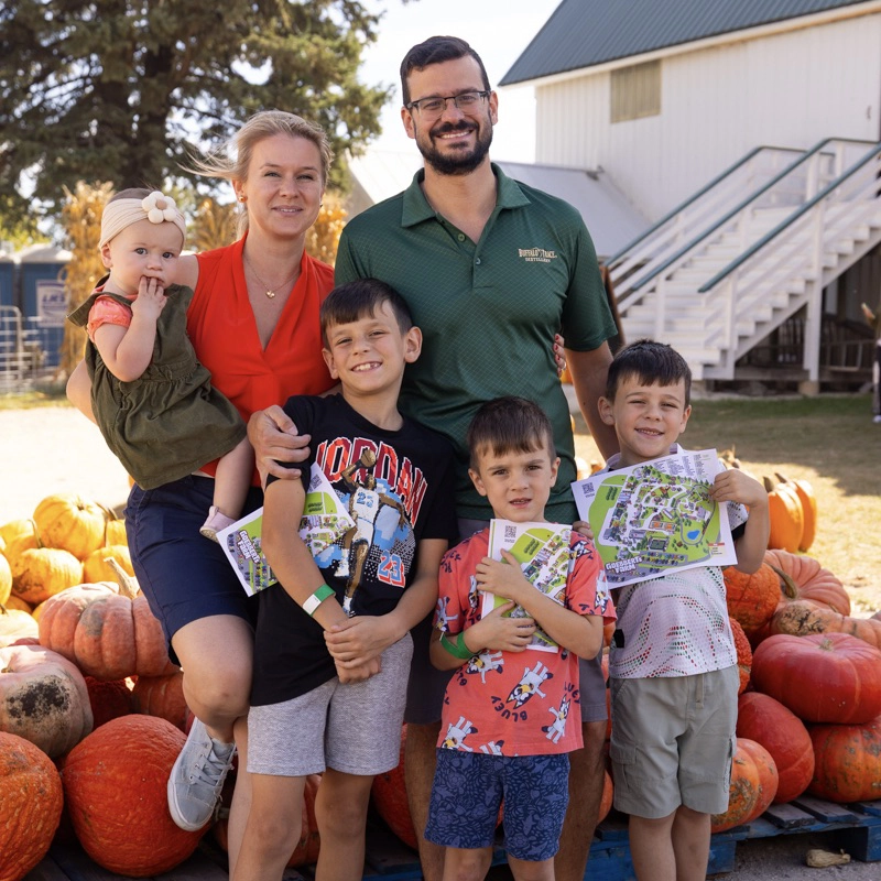 A family of five poses outdoors with pumpkins, maps in hand, in front of a white building and staircase on a sunny day.