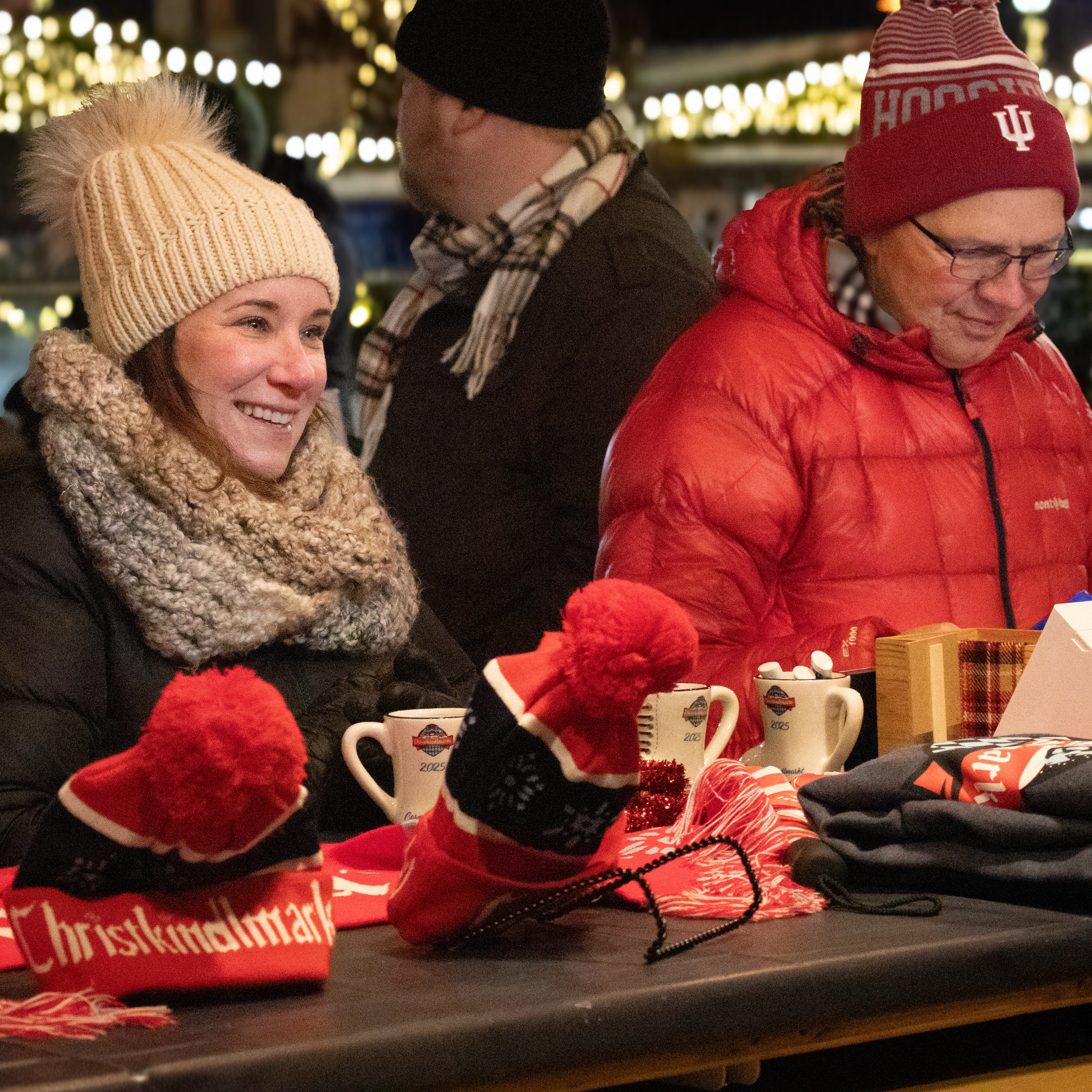 Two people in winter clothing sit at a festive outdoor booth with mugs, hats, and scarves for sale. String lights and another person are visible in the background.