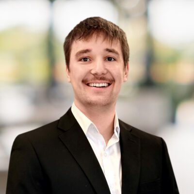 Young man with brown hair and a mustache, wearing a black suit jacket and white shirt, smiling in front of a blurred indoor background with natural light.