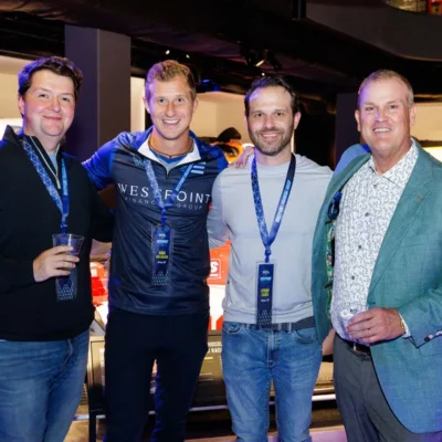 Four men stand smiling in front of race cars on display, wearing event lanyards; one holds a drink.