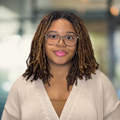 A person with glasses and shoulder-length locs wears a beige sweater and stands in front of a blurred indoor background.