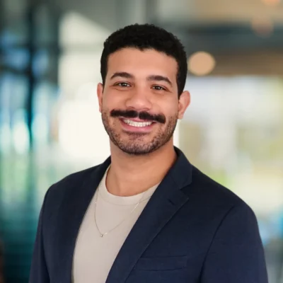A man with short curly hair, a mustache, and beard is smiling, wearing a navy blazer and beige shirt, standing in a blurred indoor setting.