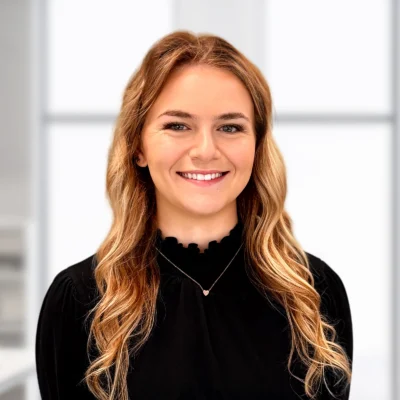 Young woman with long wavy blonde hair smiles at the camera, wearing a black top and a necklace, standing in a brightly lit modern office setting.