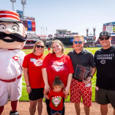 Five people and a baseball mascot stand on a baseball field, smiling for a photo. The stadium is visible in the background under a sunny sky.