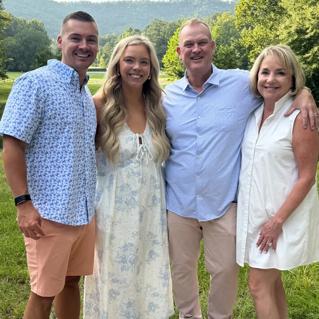 Four adults stand close together outside on grass with trees and mountains in the background, all smiling and dressed in light, summery clothing.