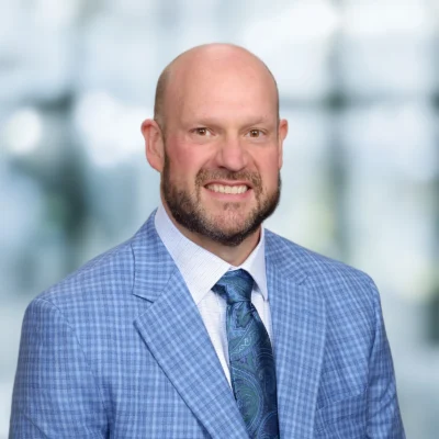 A man with a bald head and beard is wearing a blue plaid suit jacket, white shirt, and patterned blue tie, standing against a blurred background.