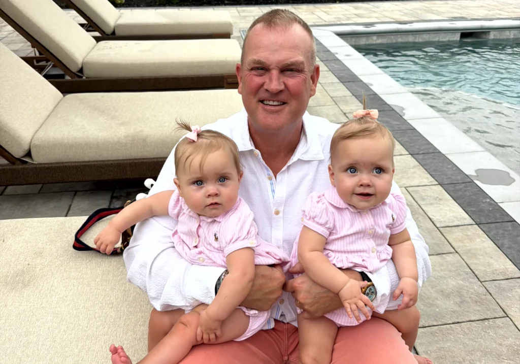 A man sits poolside on a lounge chair, holding two baby girls in matching pink outfits, one on each knee.