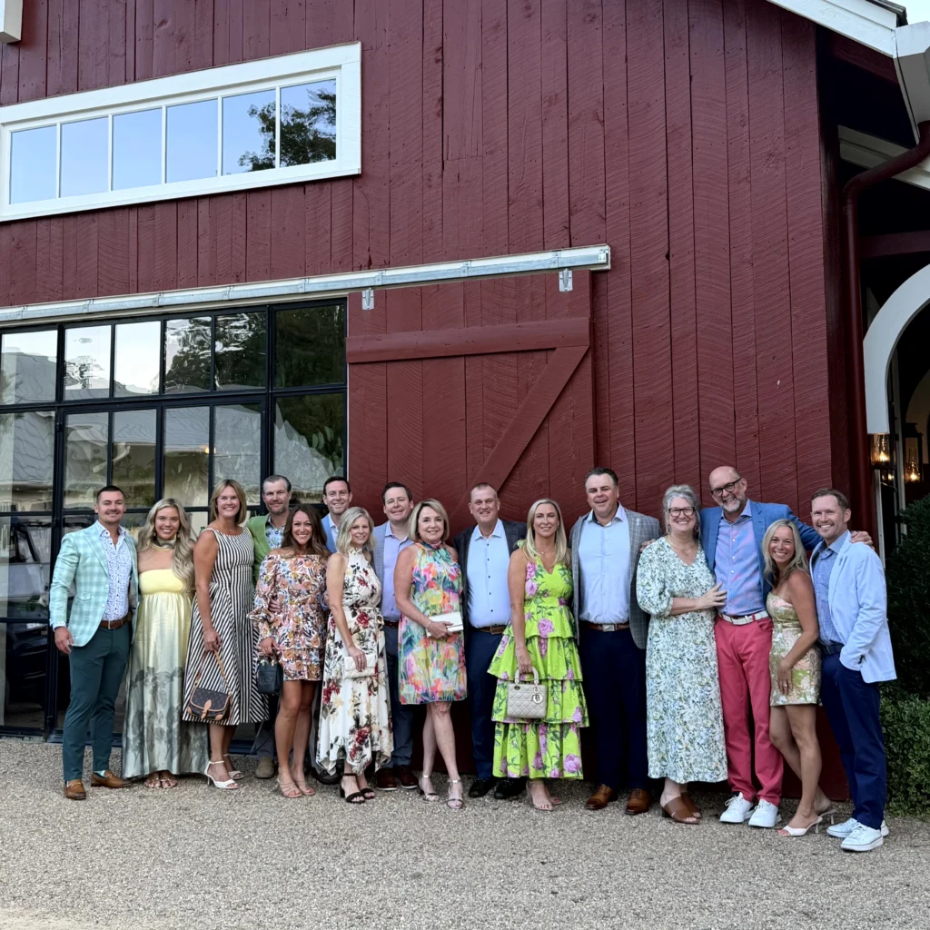 A group of eighteen adults dressed in colorful, semi-formal attire stand in a line posing outside a red wooden building.