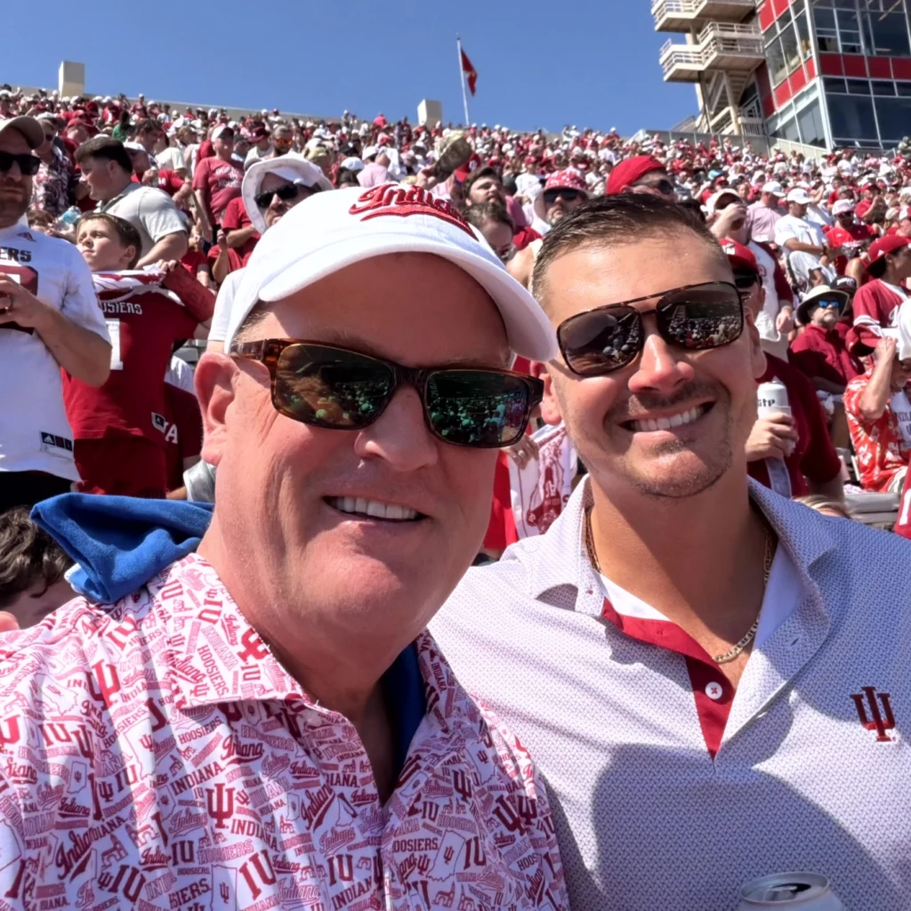 Two men wearing sunglasses and Indiana University shirts take a selfie at a crowded outdoor stadium event on a sunny day.