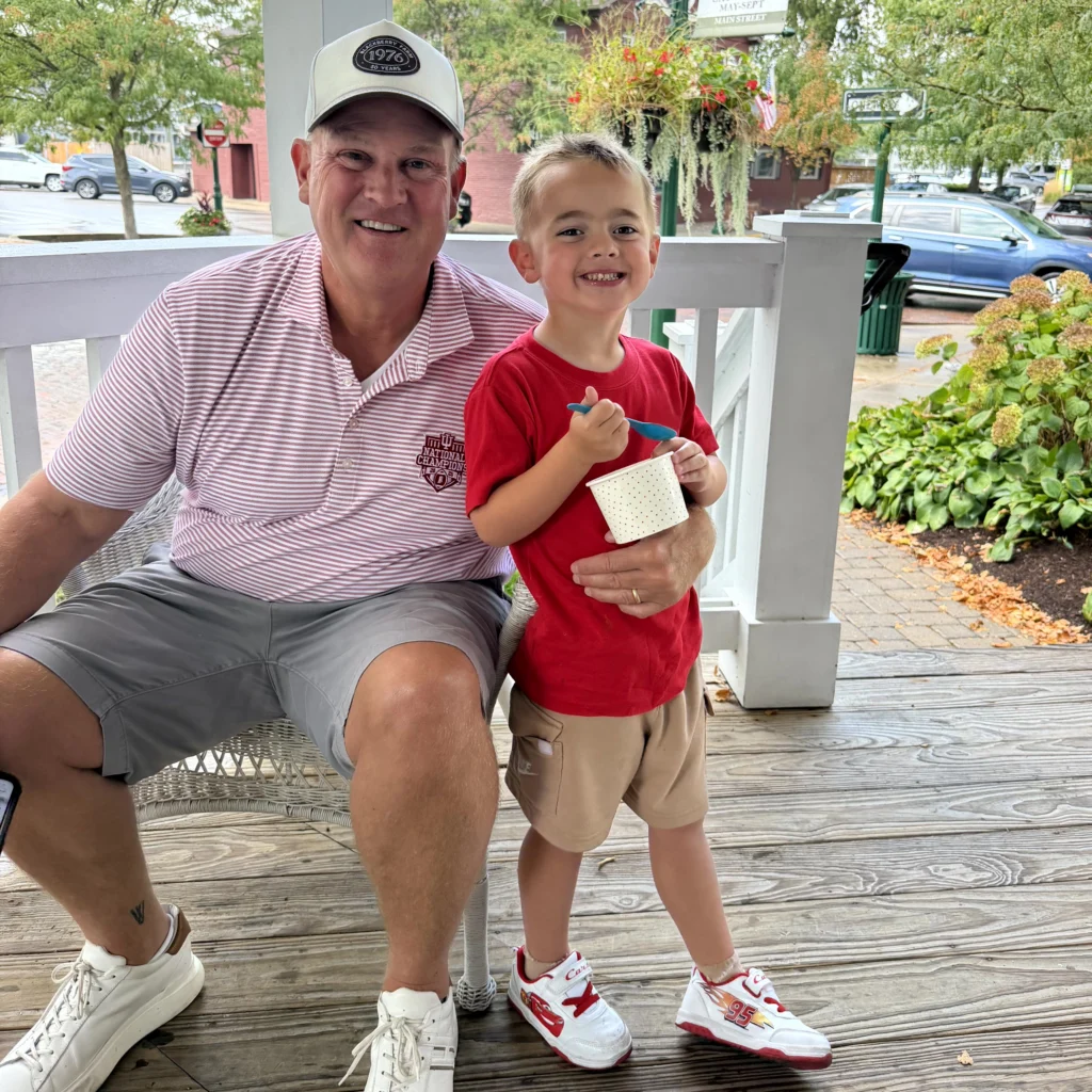 An older man and a young boy smile on a porch. The boy holds a cup of ice cream and wears a red shirt, while the man sits beside him wearing a striped shirt and shorts.