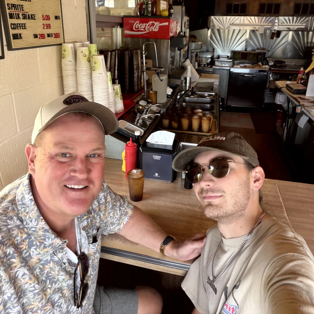 Two men sit at a diner counter with drinks, posing for a selfie. Behind them are stacked cups, condiments, and kitchen equipment. Menu board and Coca-Cola sign visible on the wall.
