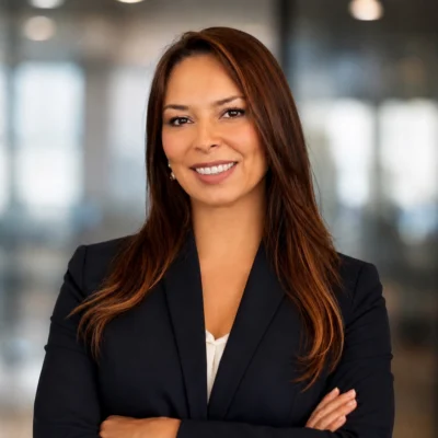 A woman with long brown hair, wearing a dark blazer and white blouse, stands with arms crossed and smiles in a modern office setting.