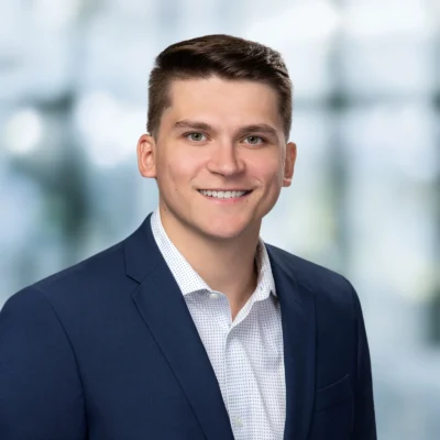 A young man wearing a navy suit jacket and white dress shirt smiles at the camera against a blurred, bright background.