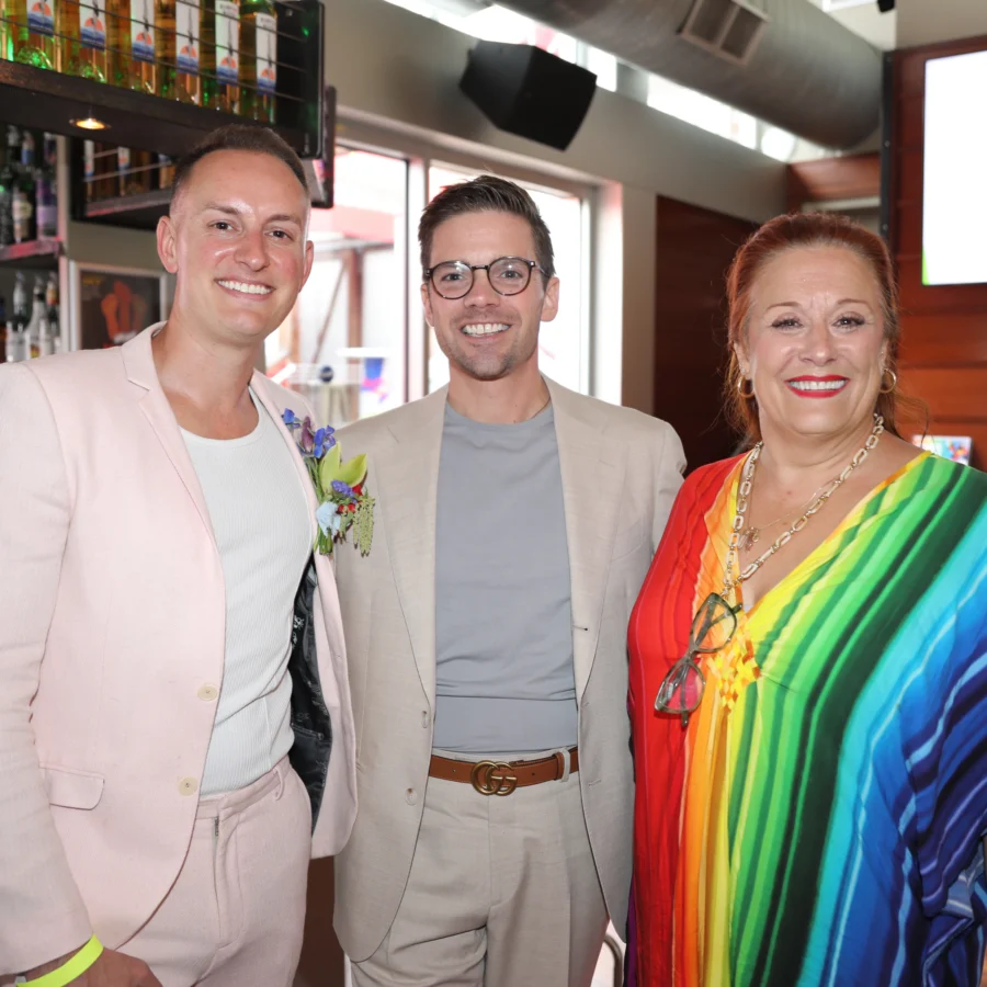 Three people smiling at the camera: a man in a pink suit, a man in a tan suit with glasses, and a woman in a rainbow-patterned top.