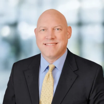Headshot of a smiling man wearing a dark suit, light blue dress shirt, and yellow patterned tie.